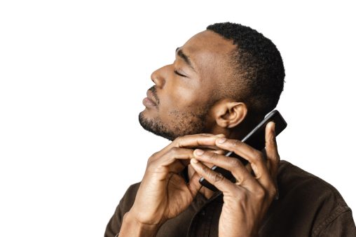 Homem de pele marrom média utiliza um celular próximo ao ouvido e com os olhos fechados. Ele possui cabelos crespos, pretos e curtos e barba rala. Veste uma camisa marrom.